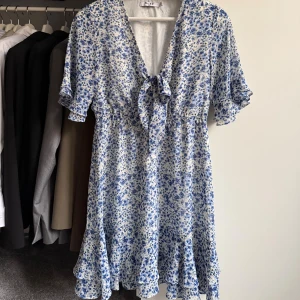 Somrig klänning - 🤍Blommig klänning i stretchigt material från NA-KD med v-ringning och knyt framtill. Klänningen har volangdetaljer vid ärmslut och fåll, samt korta ärmar 🤍