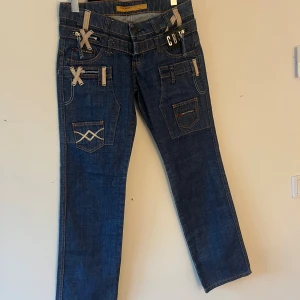 Blå jeans från Cipo & Baxx med detaljer - Snygga blå jeans från Cipo & Baxx med raka ben och coola beige snörningsdetaljer samt broderade fickor både fram och bak. Jeansens design har flera fickor, kontrastsömmar och unika detaljer som ger en streetig vibe. Perfekt för dig som gillar statement-plagg.