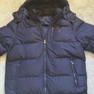 Mörkblå pufferjacka från Polo Ralph Lauren - Säljer en mörkblå pufferjacka från Polo Ralph Lauren med huva och flera dragkedjor. Jackan har det klassiska röda Polo-logot broderat på bröstet och är tillverkad i syntetmaterial. Perfekt för kalla dagar med sin tjocka och quiltade design. Använd en vinter så skicket är 9/10