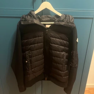 Svart cardigan från Moncler - Svart cardigan från Moncler med stickade ärmar och quiltad front. Cardigan har huva, dragkedja framtill och praktiska fickor med knapp. Snygg och sportig look med diskret Moncler-logga på ärmen. Perfekt för kyliga dagar. Den är i L men skulle verkligen säga att denna passar M lika bra, själv är jag M och har denna. Ny pris runt 9000 på denna. Denna är äkta. 
