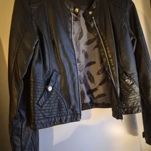 Svart skinnjacka med coola detaljer - Snygg svart skinnjacka med quiltade detaljer på axlar och nedre delen, samt silverfärgade knappar och dragkedja framtill. Jackan har lång ärm och en stilren, lite kortare passform.