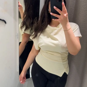 Ljusgul omlott topp med kort ärm - Säljer en ljusgul omlott topp med korta ärmar och snygg drapering över midjan. Toppen har en asymmetrisk nederkant som ger en cool touch och är tillverkad i mjukt, stretchigt material. Perfekt för dig som gillar stilrena och trendiga plagg.