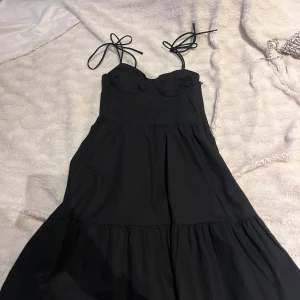 Svart klänning med knytband - Svart klänning med smala axelband som knyts på axlarna och markerad byst. Klänningen har en lätt utställd kjol och smock i ryggen för extra komfort. Perfekt för en trendig och stilren look.