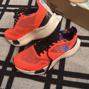 The North Face Summit Series sneakers - Säljer ett par snygga The North Face Summit Series sneakers i stark orange med svarta detaljer och blått mönstrat innerfoder. Skorna har mesh-ovandel, svart snörning och en kraftig vit plattformssula med svarta inslag. Perfekta för dig som gillar sportig stil och coola färger.