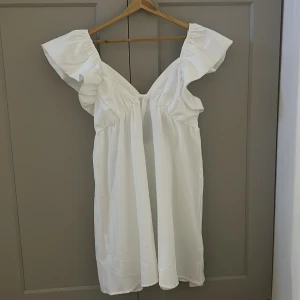 Vit kortklänning med volangärmar - Superfin vit kortklänning med breda volangärmar och v-ringning. Klänningen har en snygg knytning i ryggen och öppen detalj baktill. Luftig och lätt i materialet, perfekt för varma dagar. Passformen är normal och modellen är feminin och trendig.