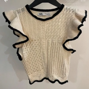 Stickad topp med volang från Zara - Säljer en krämvit stickad topp från Zara med svarta kanter och volangdetaljer längs ärmhålen. Toppen har ett vackert stickat mönster och rund halsringning. Perfekt för att lyfta en outfit!