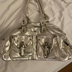 Guldskimrig handväska från Xti med detaljer - Guldskimrig från Xti med två stora fickor framtill. Väskan har rejäla handtag och dragkedja upptill. Insidan är rymlig med extra fack för småsaker. Tappat en knapp på axelremmen