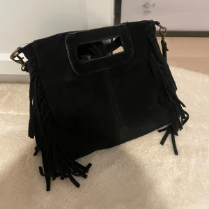 Svart mocka handväska med fransar - Cool svart handväska i äkta mocka från Borse in Pelle, tillverkad i Italien. Väskan har rektangulära handtag, avtagbar axelrem och snygga fransdetaljer på sidorna. Perfekt storlek för det viktigaste och ger en edgy vibe till din outfit. 