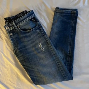 Blå Replay  - Säljer ett par snygga Replay jeans i blå med slitningar och lätt distressed detaljer. Slim fit-modell med klassiska fem fickor och dragkedja. Perfekt för dig som gillar en modern och stilren look.
