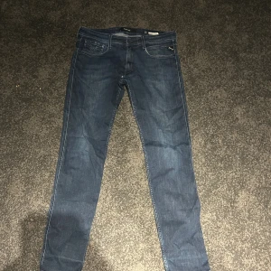 Replay Anbass mörkblå jeans herr - Prisa kan diskuteras. Snygga mörkblå Replay Anbass jeans med klassisk femficksdesign och diskreta slitningar. Slim passform och raka ben, tillverkade i mjukt jeansmaterial med stretch för extra komfort. Perfekt för dig som gillar stilrena och moderna jeans.