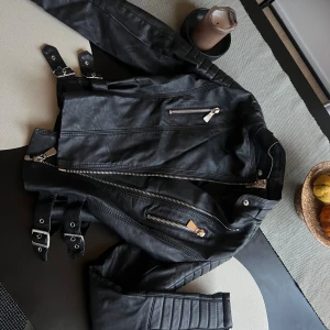 Svart skinnjacka med dragkedjor - Storlek 38 men sitter som en 34✨ Säljer en svart bikerinspirerad skinnjacka med silvriga dragkedjor och spännen i midjan. Jackan har quiltade detaljer på ärmarna och axlarna för en extra cool look. Perfekt för dig som vill ha en edgy stil.