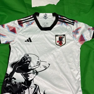 Japan fotbollströja Adidas anime S - Japan fotbollströja från Adidas i vitt med svarta detaljer och färgglada grafiska mönster på ärmarna. Tryck med anime-figur både fram och bak. Broderad Japan-logga på bröstet och japanska flaggan i nacken. Tillverkad i lätt och glansigt material.Liknande modell som originalet!