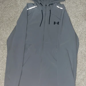 Grå vindjacka från Under Armour -  Jackan har dragkedja framtill, reflekterande detaljer på axlarna och logga på bröstet. Perfekt för träning eller bara för en jacka. Skriv om ni har frågor!