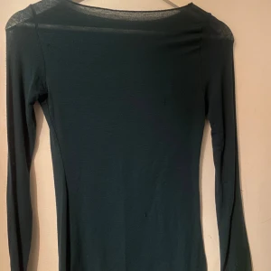 Mörkgrön långärmad tunn topp från intimissmi. - En enkel och stilren mörkgrön långärmad topp i tunn och lätt genomskinlig polyester. Modellen är figurnära med rundad halsringning och raka ärmar. Perfekt att styla med jeans eller kjol för en clean look.