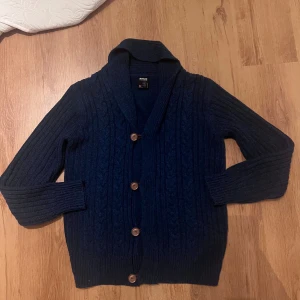 Mörkblå stickad cardigan från Smog - Säljer en mörkblå stickad kofta från Smog i storlek L. Koftan har snyggt kabelstickat mönster, bred krage och träknappar framtill. Långa ärmar och ribbade muddar ger en chill och klassisk vibe. Perfekt för dig som gillar stilren och mysig look.