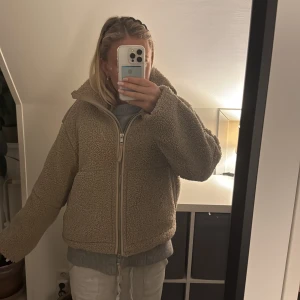 Beige teddyjacka med dragkedja - Säljer en beige teddyjacka med hög krage och två stora fickor framtill. Jackan har en grov dragkedja hela vägen och är riktigt mjuk och fluffig. Passformen är loose och den är perfekt för kalla dagar. Jackan har långa ärmar och en chill, oversized vibe.