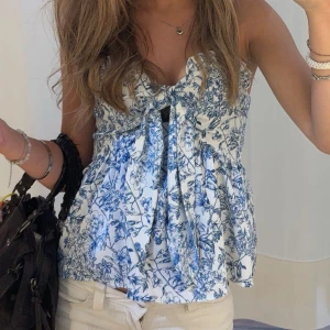 Blommig vit och blå linnetopp - Supersöt linnetopp i vitt med blått blommönster. Toppen har knytning framtill, smockad rygg och är ärmlös. Perfekt för varma dagar och ger en fräsch sommarkänsla med sin luftiga passform och romantiska vibe.