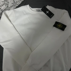 Vit sweatshirt från Stone Island - Vit sweatshirt från Stone Island med klassisk rund hals och ribbade muddar. Tröjan har den ikoniska Stone Island-loggan på vänster ärm, vilket ger en snygg streetwear-vibe. Materialet är mjukt och känns som bomull, perfekt för dig som gillar stilrena och cleana plagg.