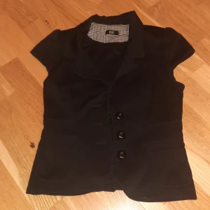 Svart väst med knappar och fickor - Stilren svart väst från F&F med korta ärmar, tre stora knappar framtill och två fickor. Super fin pris går att diskutera (lite dåligt kamerakvalitet)