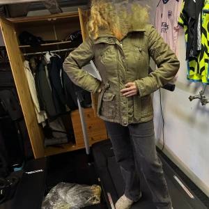 Olivgrön jacka från edc, storlek M - Snygg olivgrön jacka från edc med markerad midja och flera fickor framtill. Jackan har quiltade detaljer på insidan, knappar och dragkedja framtill samt hög krage. Perfekt för dig som vill ha en stilren och praktisk jacka med coola detaljer.