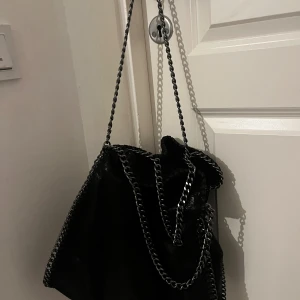 Svart axelväska med kedjedetaljer - Snygg svart axelväska med glansig finish och grova kedjor som axelrem. Väskan har en mjuk, slouchy form och insida med dragkedjefack för småsaker. Perfekt för dig som gillar en edgy stil och vill ha plats för allt viktigt. Helt oanvänd