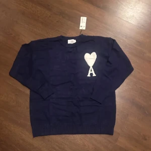 Marinblå stickad tröja från Ami - Snygg marinblå stickad tröja från Ami Alexandre Mattiussi med vit hjärtapplikation och bokstaven A på bröstet. Tröjan har rund hals, ribbade muddar och lång ärm. Perfekt för dig som gillar stilrena detaljer och vill ha något som sticker ut.