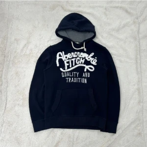 Mörkblå hoodie Abercrombie & Fitch S - Snygg mörkblå hoodie från Abercrombie & Fitch i storlek small. Tröjan har stor vit broderad logga framtill, snörning vid huvan och en klassisk känguruficka. Insidan är mjuk och gråmelerad, perfekt för chill dagar. Passar dig som gillar streetwear och avslappnad stil.