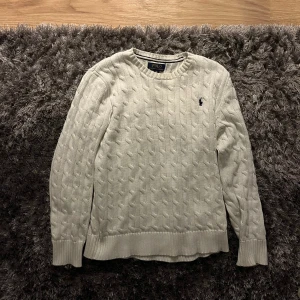 Vit kabelstickad tröja Polo Ralph Lauren - Kabelstickad vit tröja från Polo Ralph Lauren med klassisk rund halsringning och broderad logga på bröstet. Tröjan har långa ärmar och ribbade muddar för en snygg passform. Perfekt för dig som gillar stilren och tidlös design.