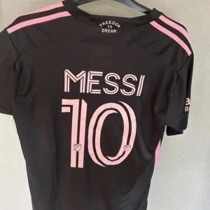 Inter Miami Messi matchtröja svart/rosa - Snygg Inter Miami fotbollströja med Messi 10-tryck på ryggen. Tröjan är svart med rosa detaljer, klubbmärke och Adidas-logga framtill. Tillverkad i lätt och ventilerande material, perfekt för fotboll eller att bära som supporter.