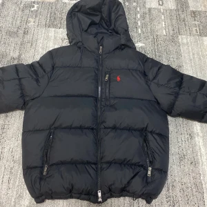 Svart pufferjacka från Polo Ralph Lauren - Svart pufferjacka från Polo Ralph Lauren med huva och röd logga på bröstet. Jackan har flera dragkedjor, inklusive två sidofickor och en innerficka. Materialet är syntet och jackan är quiltad för extra värme. Perfekt för kalla dagar. Perfekt för storlek M och L