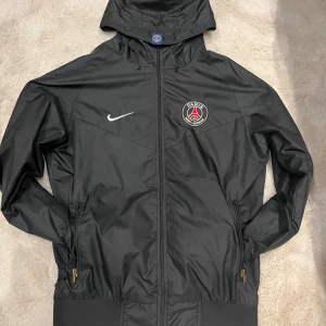 Svart PSG vindjacka från Nike - Svart vindjacka från Nike x Paris Saint-Germain med broderad PSG-logga och Nike Swoosh på bröstet. Jackan har huva, hel dragkedja och två fickor med dragkedja. Lätt och sportig design i syntetmaterial, perfekt för fotbollsfans.