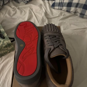 Gråa Louboutin sneakers med nitar - Säljer ett par gråa sneakers från Christian Louboutin i mocka med ikonisk röd sula och silverfärgade nitar över tån. Skorna har snörning och en rund tå, samt beige innersida för extra komfort. Perfekta för dig som vill sticka ut med en edgy stil.