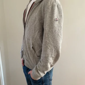 Grå Moncler hoodie med dragkedja - Snygg grå hoodie från Moncler i storlek L (ungdom) passar folk runt 170-176.  Tröjan har huva,  dragkedja med röd och blå detalj.  Finns en liten fläck på tröjan men går nog bort i tvätten bara 