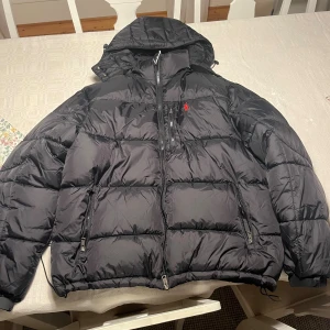 Svart pufferjacka Polo Ralph Lauren XL - Svart pufferjacka från Polo Ralph Lauren i storlek XL, men passar L. Jackan har huva, flera dragkedjor och den klassiska röda Polo-loggan broderad på bröstet. Materialet är syntet och jackan är perfekt för kalla dagar. Snygg och stilren design med quiltade sektioner.