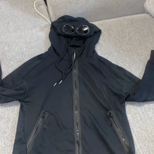 Svart C.P. Company goggle jacka - Säljer en svart goggle jacka från C.P. Company med huva och inbyggda glasögon. Jackan har dragkedja framtill, två snedställda fickor med dragkedja och justerbar snörning vid huvan. Perfekt för dig som gillar streetwear och vill sticka ut.