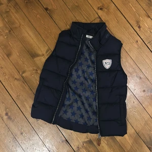 Marinblå dunväst från H&M  - Snygg marinblå dunväst från H&M med quiltad design och dragkedja framtill. Insidan har ett coolt stjärnmönster i blått och grått. På bröstet sitter en patch med texten 'No 1'. Perfekt för lager-på-lager och streetstyle.