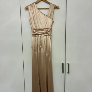 Champagnefärgad långklänning i satin - Elegant långklänning i skimrande champagnefärgad satin med oneshoulder-design. Klänningen har draperad midja och slät, figurnära siluett som ger en lyxig känsla. Perfekt för dig som vill sticka ut med en stilren och modern look.