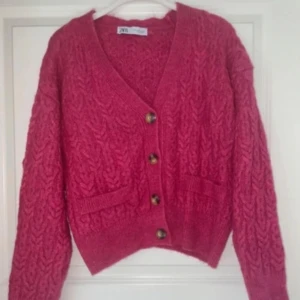 Rosa stickad kofta från Zara - Superfin rosa stickad kofta från Zara med snygg kabelstickad struktur, v-ringning och fyra knappar framtill. Koftan har två små fickor och långa ärmar. Perfekt för lager-på-lager och ger en mysig vibe till din outfit.