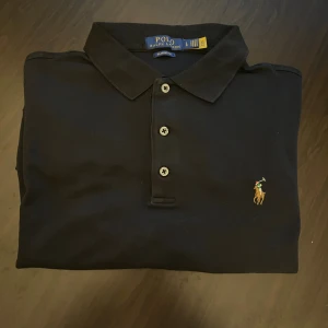 Svart lång ärmar pikétröja från Polo Ralph Lauren - Klassisk svart pikétröja från Polo Ralph Lauren i bomull med broderad logga på bröstet. Tröjan har krage, tre knappar framtill och en clean, tidlös look. Perfekt för dig som gillar stilrena och ikoniska plagg.