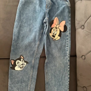 Blå jeans med Disney-tryck - Unika blå jeans med coola tryck av Minnie Mouse och Figaro på benen. Jeansen har en klassisk rak passform och är gjorda i jeansmaterial. Perfekt för dig som vill sticka ut med lite Disney-vibe i din outfit.