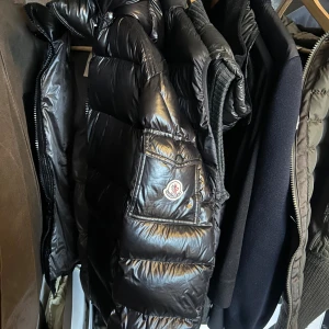 Moncler Maya  - Säljer en svart, glansig pufferjacka från Moncler med huva och klassisk logga på armen. Jackan har dragkedja framtill och är vadderad för extra värme. Insidan har ett unikt serietryck. Perfekt för kalla dagar när du vill hålla stilen. Kvitto mer bilder på tags kom pm. Två små håll på armen inget som läggs märke till dragkedjan nertill är sönder men alla delar finns till att fixa den igen passar S/M