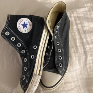Svarta Converse All Star high-tops - Klassiska svarta Converse All Star high-tops med vit tå och sula. Skorna har rund tå, vita kontrastsömmar och metallöljetter för snörning. Tillverkade i canvas med gummisula och ikonisk logga på sidan. Perfekta för en avslappnad och cool stil.