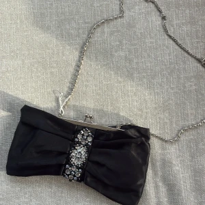 Svart kuvertväska med strassdetaljer - Elegant svart kuvertväska i satin med silverfärgad kedja och glittrande strassdetaljer framtill. Väskan har en klassisk metallknäppning och en snygg rosettformad design som gör den extra festlig. Perfekt för att lyfta din outfit till nästa nivå.