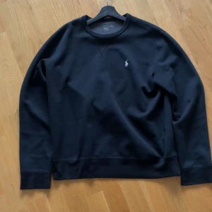 Svart sweatshirt från Ralph Lauren - Klassisk svart sweatshirt från Ralph Lauren med rund halsringning och diskret broderad logga på bröstet. Tröjan har lång ärm och är tillverkad i mjuk bomull, perfekt för en stilren och avslappnad look.