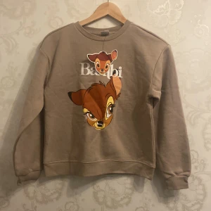 Bambi Tröja - Superfin beige sweatshirt med stort Bambi-tryck på framsidan och vit text. Tröjan har rund hals, långa ärmar och ribbade muddar vid ärmslut och nederkant. Perfekt för dig som älskar Disney och vill ha en mysig och snygg tröja. 152cm tröja. !OBS Skickas ej, finns i Rissne att hämta.