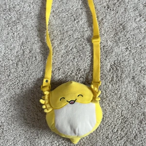 Gul Squishmallow-axelväska djurmotiv-NY! ❣️ - Supergullig Squishmallow axelväska i mjuk plysch med ett gult djurmotiv och justerbar rem. Väskan har ett glatt ansikte framtill och en dragkedja på baksidan med bruna detaljer. ❣️