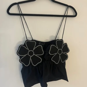 Svart croppad linnetopp med blommor - Supersöt svart croppad linnetopp från ZARA med tunna axelband och två stora blommor framtill, dekorerade med glittriga pärlor längs kanterna. Toppen har en luftig passform och är perfekt för att sticka ut med en unik look.