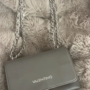 Grå axelväska från Valentino - Snygg grå axelväska från Valentino med silverfärgad kedjerem och stilren design. Väskan är i slätt skinn och har flera fack inuti, inklusive ett med dragkedja. Perfekt för dig som vill ha en trendig och praktisk väska med exklusiv känsla.