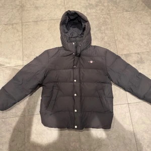 Svart pufferjacka från GANT - Svart pufferjacka från GANT med huva och snyggt  logomärke på bröstet. Jackan har dragkedja och knappar framtill, två fickor och justerbar snörning nedtill. Perfekt för kalla dagar och har en clean look.