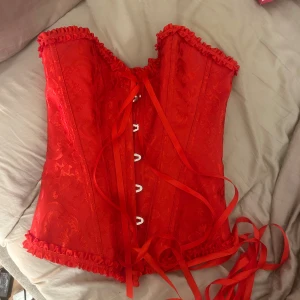Röd korsett med snörning och volang - Säljer en intensivt röd korsett med snygg snörning framtill och dekorativa volanger längs kanterna. Korsetten har ett subtilt mönster i tyget och är perfekt för dig som vill sticka ut. Materialet känns glansigt och lyxigt, och snörningen gör att du kan justera passformen.